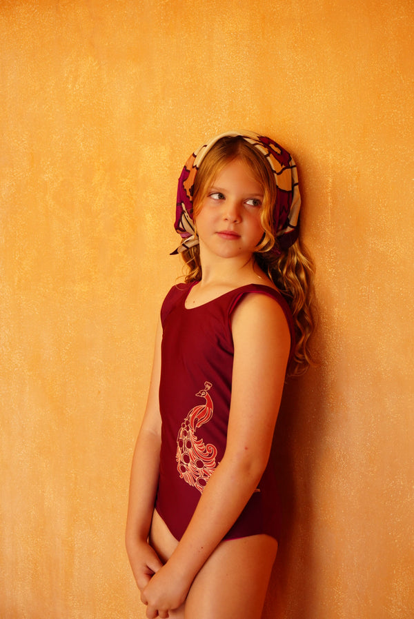 A young girl wears a wine-colored one-piece swimsuit with a fuchsia peacock design on the front.