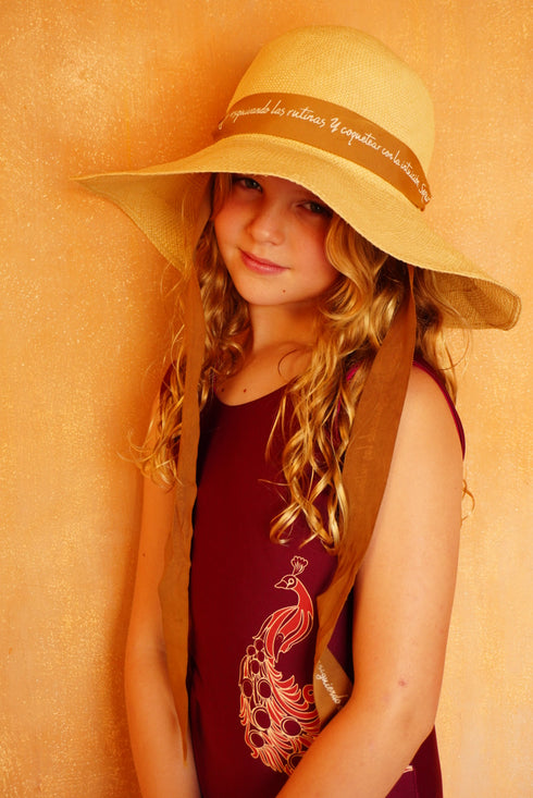 Young girl wearing a wine-colored one-piece swimsuit with a peacock graphic and a wide-brimmed hat.