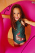 A young girl wears a green one-shoulder swimsuit featuring a red and white peacock graphic.